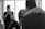 A black and white photograph captures an emotional moment. A woman in the background is wiping tears with a tissue, while a man with a beard, dressed in a suit, sits in the foreground. In the blurry foreground, two people embrace closely, suggesting an intimate moment, possibly a dance or embrace.