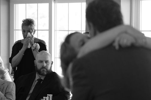 A black and white photograph captures an emotional moment. A woman in the background is wiping tears with a tissue, while a man with a beard, dressed in a suit, sits in the foreground. In the blurry foreground, two people embrace closely, suggesting an intimate moment, possibly a dance or embrace.