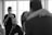 A black and white photograph captures an emotional moment. A woman in the background is wiping tears with a tissue, while a man with a beard, dressed in a suit, sits in the foreground. In the blurry foreground, two people embrace closely, suggesting an intimate moment, possibly a dance or embrace.