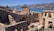 Ancient stone ruins overlook a calm blue sea with mountains in the background. The scene includes a small boat on the water and part of a modern building to the right. Vegetation grows sporadically among the weathered stone structures, and the clear sky above suggests a sunny day.