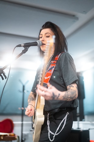 A musician with tattoos is playing an electric guitar and singing into a microphone. The background is softly lit, and the performer wears a black t-shirt and pants with chains. The guitar strap is adorned with red and white designs.