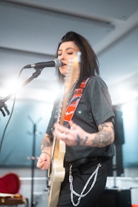 A musician with tattoos is playing an electric guitar and singing into a microphone. The background is softly lit, and the performer wears a black t-shirt and pants with chains. The guitar strap is adorned with red and white designs.