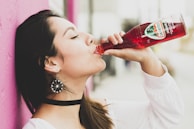 A woman with long dark hair, adorned with decorative earrings and wearing a white top, enjoys a drink from a red bottle labeled Jarritos. She stands against a bright pink wall, with a background that appears softly blurred.