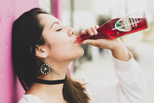 A woman with long dark hair, adorned with decorative earrings and wearing a white top, enjoys a drink from a red bottle labeled Jarritos. She stands against a bright pink wall, with a background that appears softly blurred.