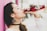 A woman with long dark hair, adorned with decorative earrings and wearing a white top, enjoys a drink from a red bottle labeled Jarritos. She stands against a bright pink wall, with a background that appears softly blurred.