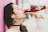 A woman with long dark hair, adorned with decorative earrings and wearing a white top, enjoys a drink from a red bottle labeled Jarritos. She stands against a bright pink wall, with a background that appears softly blurred.