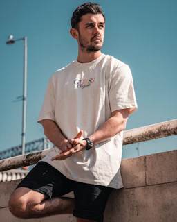 man in white crew neck t-shirt and black pants sitting on brown wooden fence during