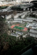 Aerial view of a residential area with several multi-story apartment buildings arranged in a semi-circular pattern. A sports complex with a green soccer field and red running track is in the center. Surrounding the area are patches of dense green forest and various roads intersecting the terrain. The landscape is largely urban with hints of green spaces.