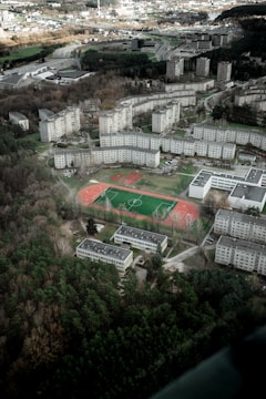 Aerial view of a residential area with several multi-story apartment buildings arranged in a semi-circular pattern. A sports complex with a green soccer field and red running track is in the center. Surrounding the area are patches of dense green forest and various roads intersecting the terrain. The landscape is largely urban with hints of green spaces.