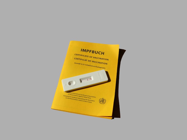 A vaccination certificate booklet is placed on a flat surface, along with a rapid test cassette lying on top. The booklet is yellow, with text indicating it as a certificate of vaccination, and bears the seal of the World Health Organization. The rapid test cassette is white, simple in design, and oriented horizontally.