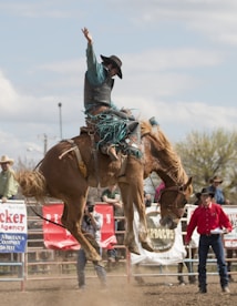 A rodeo event with a person riding a bucking horse. The rider is wearing a cowboy hat and a protective vest, holding one arm up for balance. Spectators and other participants are visible in the background, along with fences and banners.