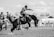 A black and white photograph depicting a rodeo scene with a cowboy riding a bucking horse. Several bystanders and other cowboys are visible in the background watching the event. The action is captured mid-motion, highlighting the dramatic movement of the horse and the concentration of the rider.