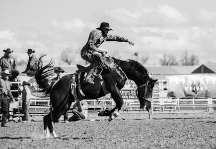 A black and white photograph depicting a rodeo scene with a cowboy riding a bucking horse. Several bystanders and other cowboys are visible in the background watching the event. The action is captured mid-motion, highlighting the dramatic movement of the horse and the concentration of the rider.