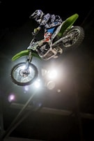 A motorcyclist performs a high jump stunt on a green dirt bike in an indoor setting with lights illuminating the scene.