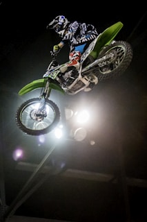A motorcyclist performs a high jump stunt on a green dirt bike in an indoor setting with lights illuminating the scene.