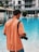 A person with a camera slung over their shoulder stands by a poolside, wearing an orange shirt and a dark cap. The setting appears to be a modern hotel or resort with lounging chairs and a building in the background.