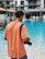 A person with a camera slung over their shoulder stands by a poolside, wearing an orange shirt and a dark cap. The setting appears to be a modern hotel or resort with lounging chairs and a building in the background.
