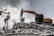 A construction excavator is engaged in demolition work, surrounded by smoke and dust. The machinery is positioned on a pile of rubble, consisting of broken concrete and twisted metal. A 'no parking' sign is displayed among the debris, and a partially destroyed building is visible in the background.