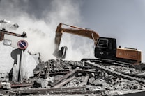 A construction excavator is engaged in demolition work, surrounded by smoke and dust. The machinery is positioned on a pile of rubble, consisting of broken concrete and twisted metal. A 'no parking' sign is displayed among the debris, and a partially destroyed building is visible in the background.