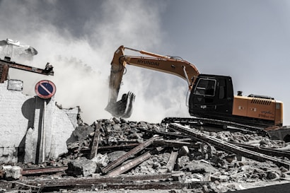 A construction excavator is engaged in demolition work, surrounded by smoke and dust. The machinery is positioned on a pile of rubble, consisting of broken concrete and twisted metal. A 'no parking' sign is displayed among the debris, and a partially destroyed building is visible in the background.