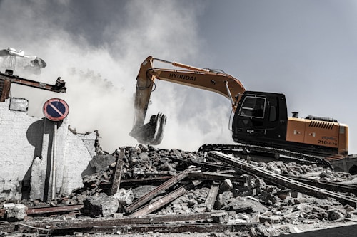 A construction excavator is engaged in demolition work, surrounded by smoke and dust. The machinery is positioned on a pile of rubble, consisting of broken concrete and twisted metal. A 'no parking' sign is displayed among the debris, and a partially destroyed building is visible in the background.