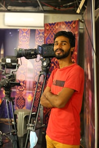 A man in a red shirt stands confidently with his arms crossed beside a professional video camera on a tripod. The setting appears to be a studio or a set, with decorative patterns on the purple and orange walls in the background. A face mask hanging from the camera suggests recent times when masks were commonly used.