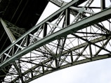A detailed view of a metallic bridge structure featuring intricate lattice work and riveted connections. The geometric pattern of the steel beams creates a complex and repetitive design against a bright white background.