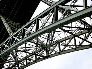 A detailed view of a metallic bridge structure featuring intricate lattice work and riveted connections. The geometric pattern of the steel beams creates a complex and repetitive design against a bright white background.