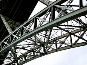 A detailed view of a metallic bridge structure featuring intricate lattice work and riveted connections. The geometric pattern of the steel beams creates a complex and repetitive design against a bright white background.