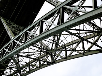A detailed view of a metallic bridge structure featuring intricate lattice work and riveted connections. The geometric pattern of the steel beams creates a complex and repetitive design against a bright white background.