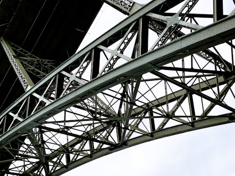 A detailed view of a metallic bridge structure featuring intricate lattice work and riveted connections. The geometric pattern of the steel beams creates a complex and repetitive design against a bright white background.