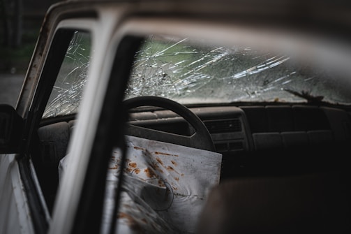 A close-up view of a car interior with a cracked windshield. The steering wheel is visible, and there is a sheet draped over the passenger seat with rust-colored stains. The dashboard is dusty, and the mood is gloomy.