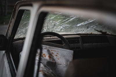 A close-up view of a car interior with a cracked windshield. The steering wheel is visible, and there is a sheet draped over the passenger seat with rust-colored stains. The dashboard is dusty, and the mood is gloomy.