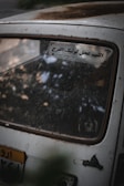 A close-up view of the rear window of an old car, covered in dust and dirt. There is a sticker with Arabic text on the window, and a portion of the rear license plate is visible.