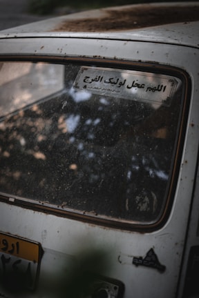 A close-up view of the rear window of an old car, covered in dust and dirt. There is a sticker with Arabic text on the window, and a portion of the rear license plate is visible.