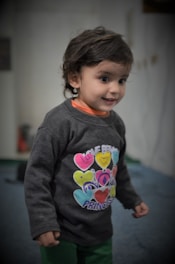 A young child with dark hair and bright eyes is wearing a colorful shirt featuring heart designs and the words 'Love Being a Princess'. The background is softly blurred, suggesting an indoor setting.
