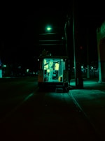 A food truck is parked on a dimly lit street at night, illuminated by a greenish light. The darkness of the surroundings contrasts with the bright interior where a person is visible inside the truck.