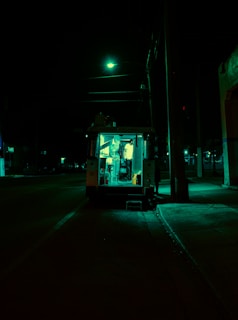 A food truck is parked on a dimly lit street at night, illuminated by a greenish light. The darkness of the surroundings contrasts with the bright interior where a person is visible inside the truck.