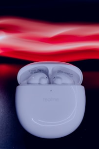 A pair of white wireless earbuds is positioned inside an open charging case with 'realme' branding on the front. The background consists of a dark surface with abstract, fluid light streaks in shades of red and white.