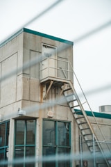 A concrete industrial building with a metal staircase leading to an elevated door marked 'Elevator Machine Room'. The structure has large glass windows with teal frames on the ground level and a metallic fence partially visible in the foreground. The overall environment looks industrial and functional.