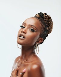 A woman with styled braided hair and hoop earrings poses gracefully with visible confidence and poise. Her makeup includes bold eyeliner and glossy lips, complemented by long, manicured nails. The background is soft and neutral, highlighting her features.