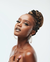 A woman with styled braided hair and hoop earrings poses gracefully with visible confidence and poise. Her makeup includes bold eyeliner and glossy lips, complemented by long, manicured nails. The background is soft and neutral, highlighting her features.