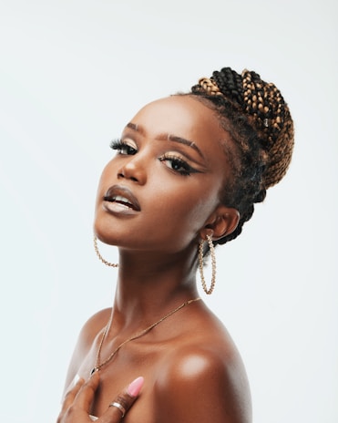 A woman with styled braided hair and hoop earrings poses gracefully with visible confidence and poise. Her makeup includes bold eyeliner and glossy lips, complemented by long, manicured nails. The background is soft and neutral, highlighting her features.