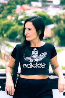 A person with long dark hair is wearing a black Adidas crop top with a floral pattern. They are standing outdoors with a blurred background of green foliage and houses. The person is leaning against a barrier, with one hand resting on it, and has a relaxed expression.