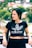 A person with long dark hair is wearing a black Adidas crop top with a floral pattern. They are standing outdoors with a blurred background of green foliage and houses. The person is leaning against a barrier, with one hand resting on it, and has a relaxed expression.