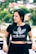 A person with long dark hair is wearing a black Adidas crop top with a floral pattern. They are standing outdoors with a blurred background of green foliage and houses. The person is leaning against a barrier, with one hand resting on it, and has a relaxed expression.