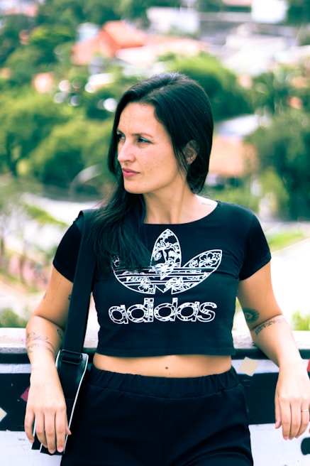 A person with long dark hair is wearing a black Adidas crop top with a floral pattern. They are standing outdoors with a blurred background of green foliage and houses. The person is leaning against a barrier, with one hand resting on it, and has a relaxed expression.