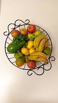 A metal fruit basket containing a variety of fruits and greens. The basket holds bananas, apples, lemons, oranges, an avocado, and some parsley. The basket has a decorative design and is placed on a white surface.