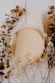 A round, gold-framed mirror is hanging by a chain surrounded by dried leaves, delicate white flowers, and ornamental grasses. The foliage is arranged symmetrically around the mirror, creating an organic, natural frame that complements the warm tones of the gold. The background is neutral, enhancing the earthy and rustic aesthetic of the arrangement.