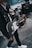 A person wearing a black leather jacket is holding a black and white electric guitar. In the background, a drummer is playing on a drum kit, and there are amplifiers visible. The setting appears to be a music studio or rehearsal space.